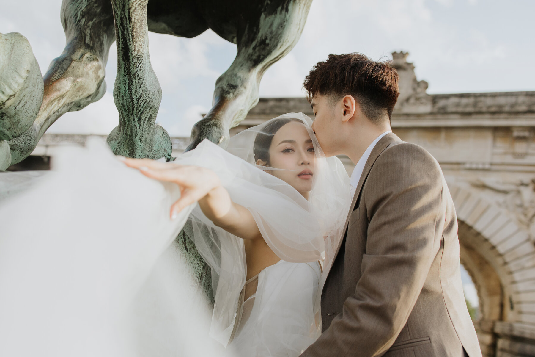 Bride reaching toward a statue, groom kissing her forehead